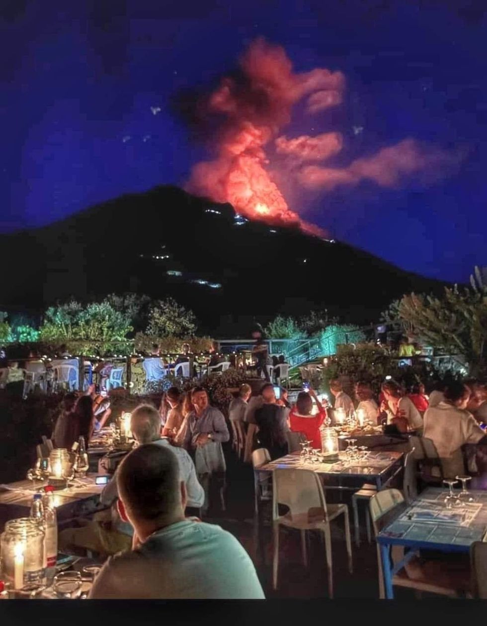 Photograph of a group of people at dinner who look up to the distant Stromboli volcano, which is lit up with its own mini-eruptions