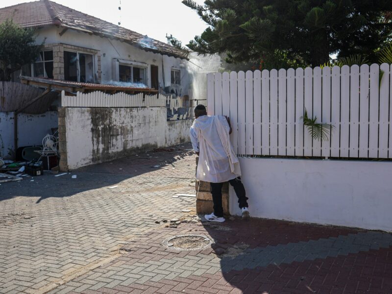 Photograph of a man peeking around a corner to inspect the damage caused by a rocket attack from Gaza.