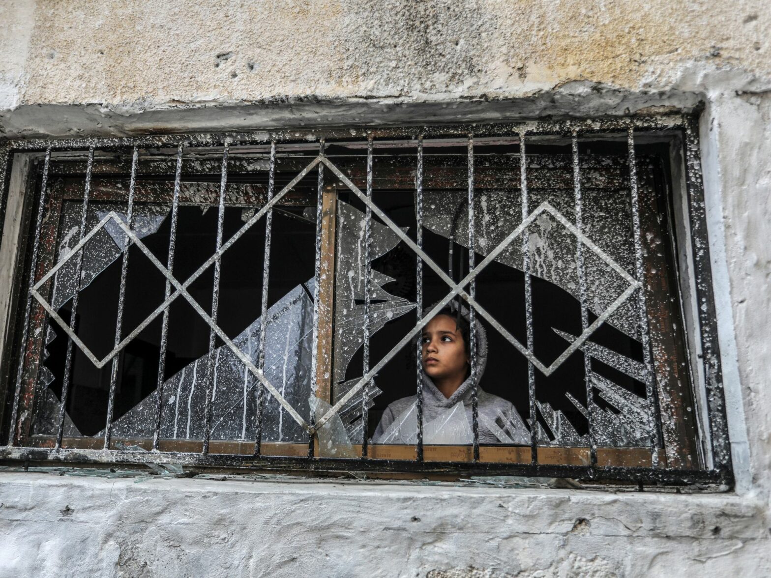 Photograph of a Palestinian child looks through a broken window of a destroyed house