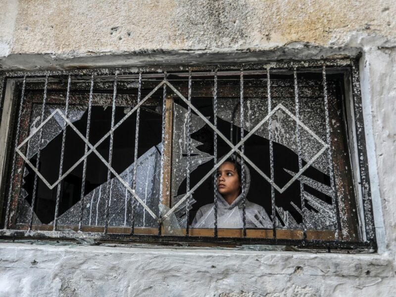 Photograph of a Palestinian child looks through a broken window of a destroyed house