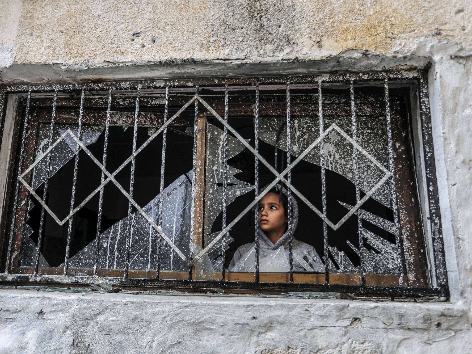 Photograph of a Palestinian child looks through a broken window of a destroyed house