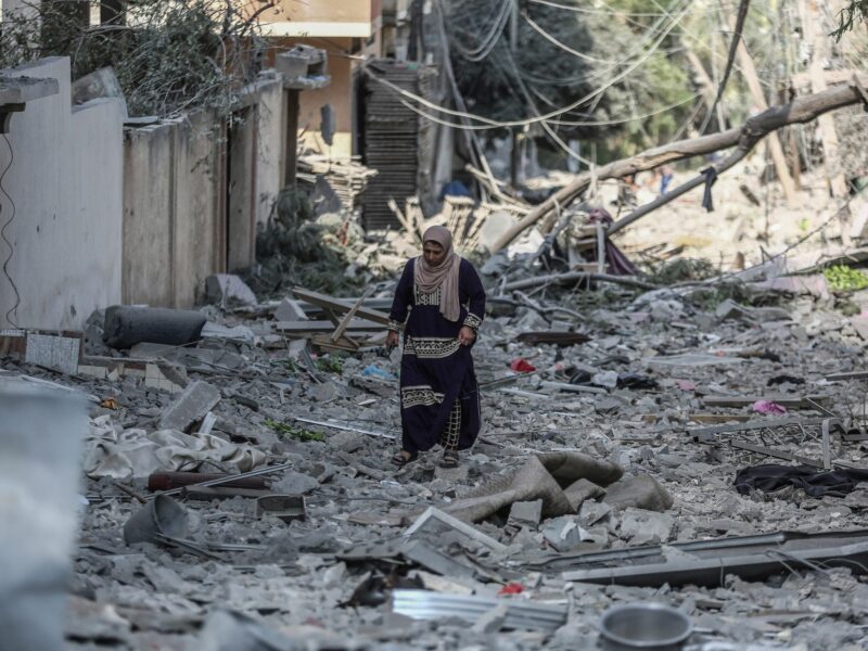 Photograph of a Palestinian woman walks through the rubble of the Karama neighborhood following an Israeli bombing.