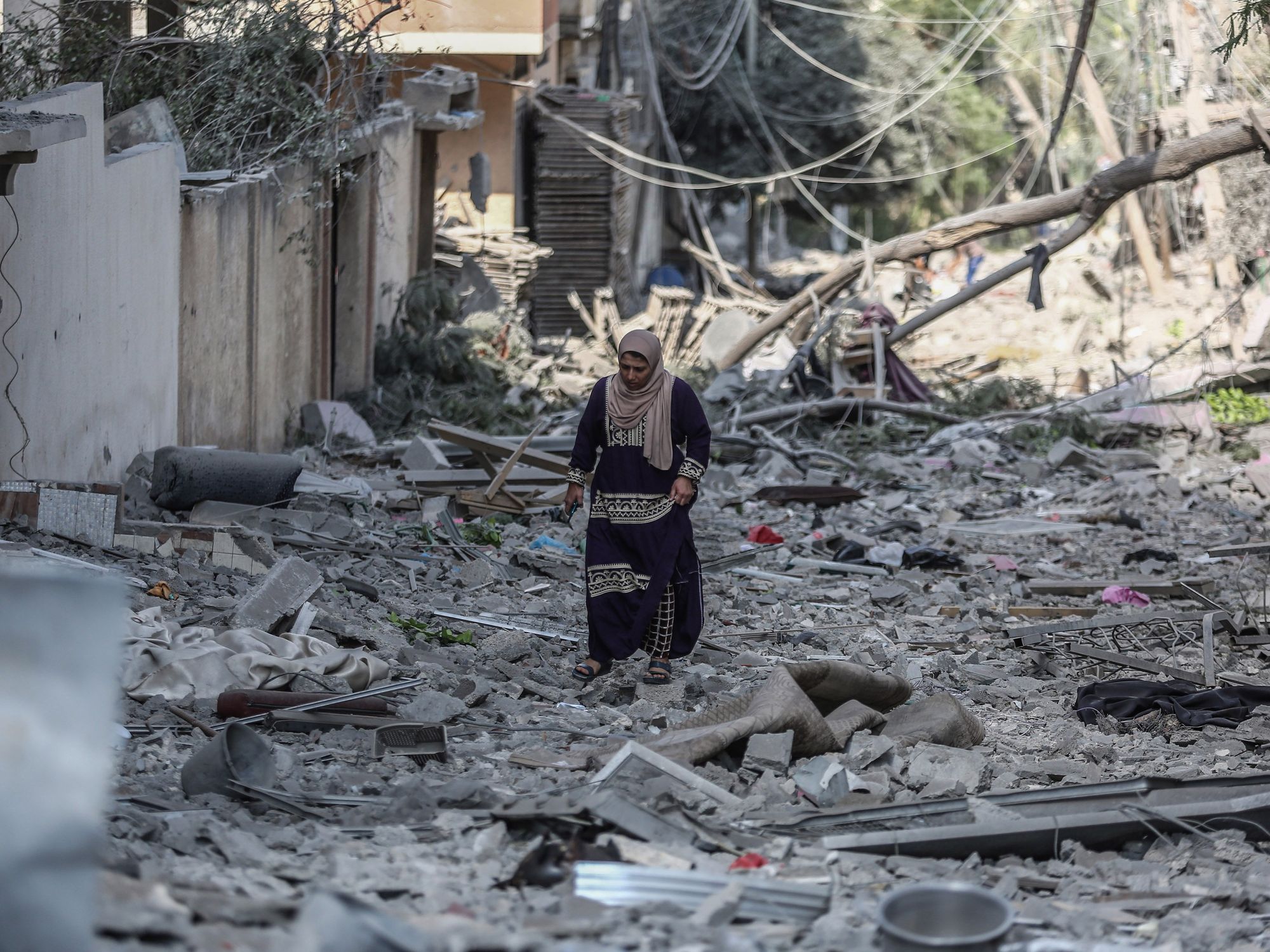 Photograph of a Palestinian woman walks through the rubble of the Karama neighborhood following an Israeli bombing.