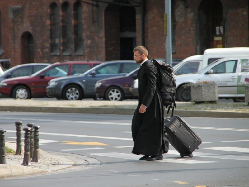 Photograph of a priest crossing the road with luggage in hand as he heads to Tumski Island, Poland.