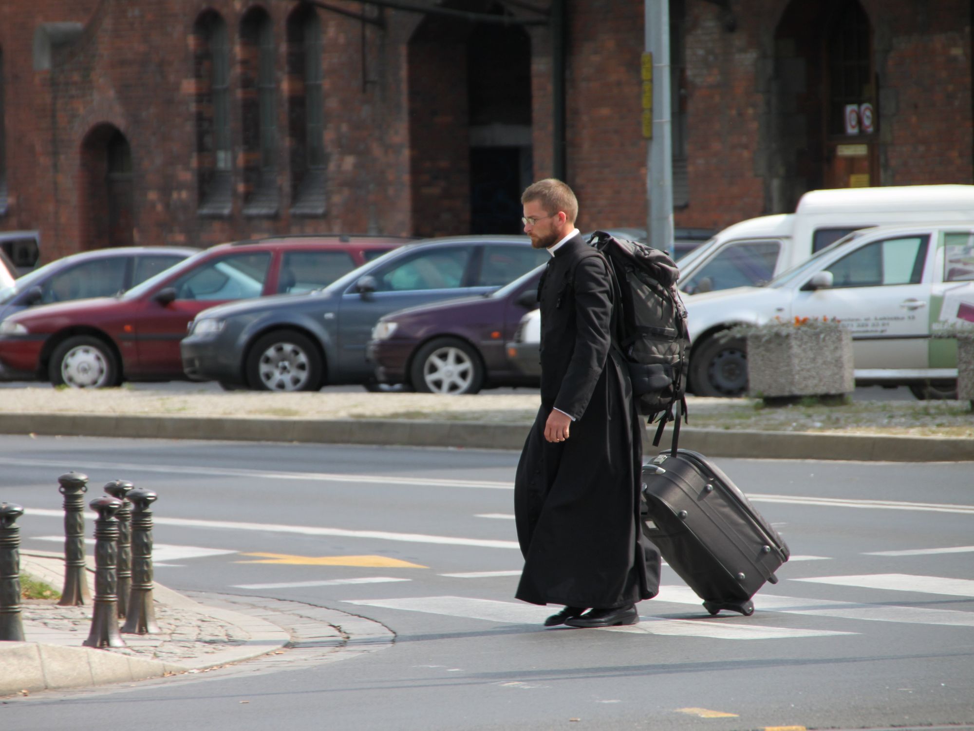 Photograph of a priest crossing the road with luggage in hand as he heads to Tumski Island, Poland.