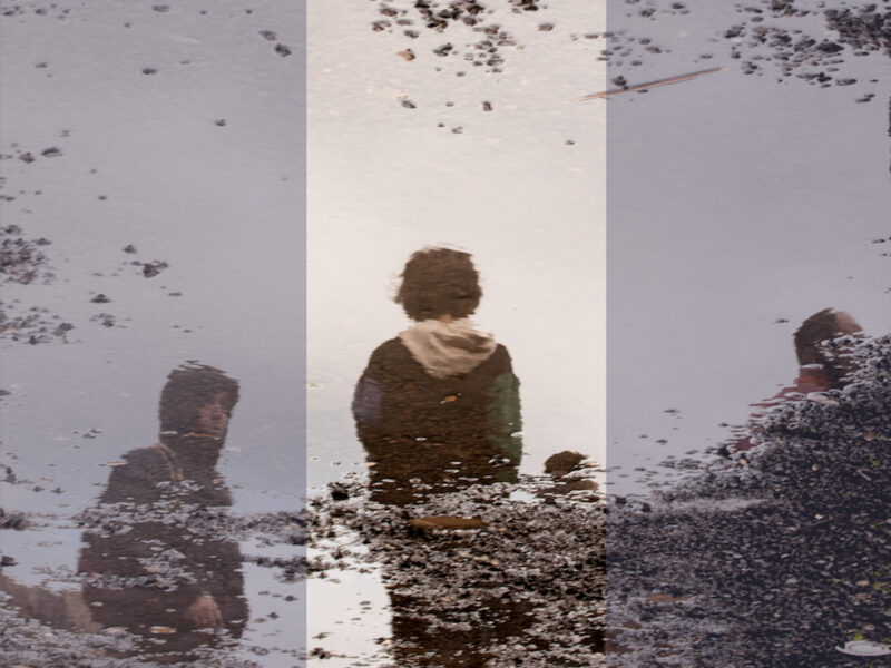 Photograph of a puddle's reflection, where children stand by the side of a gravel street