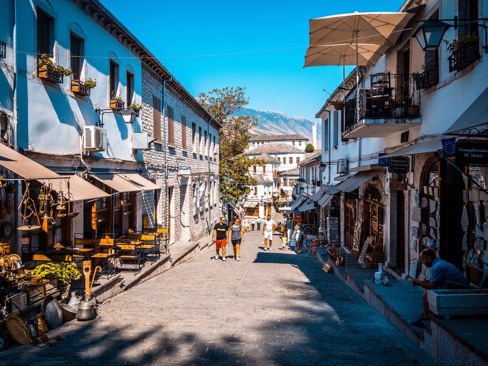 Photograph of a street in Gjirokaster, Albania. A couple of people walk down the sunny stone street, and in the distance there are mountains.