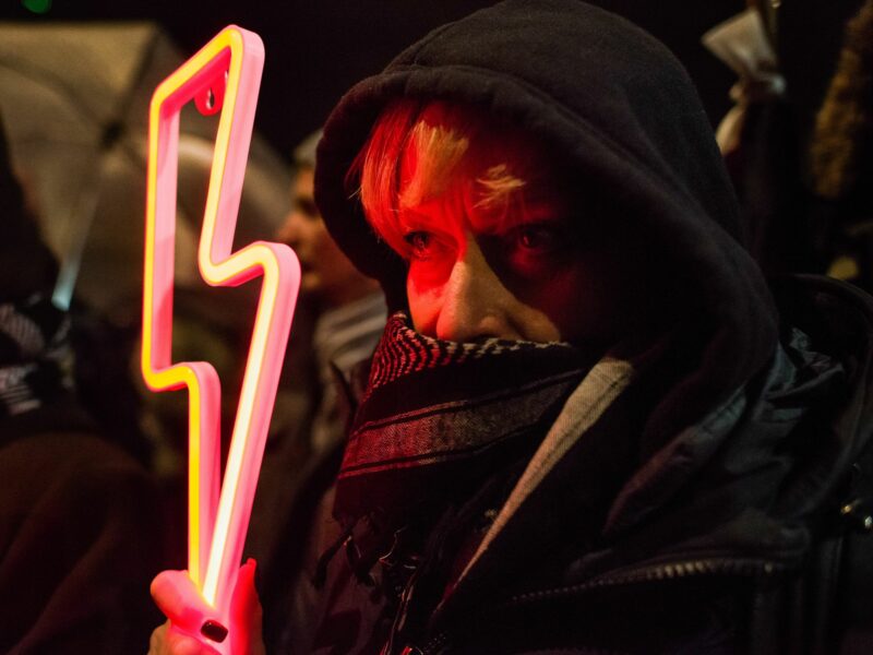 Photograph of a woman with her lower face covered holding a red lightning bolt - the symbol of the Women's Strike - during the demonstration outside Kaczynski's house.