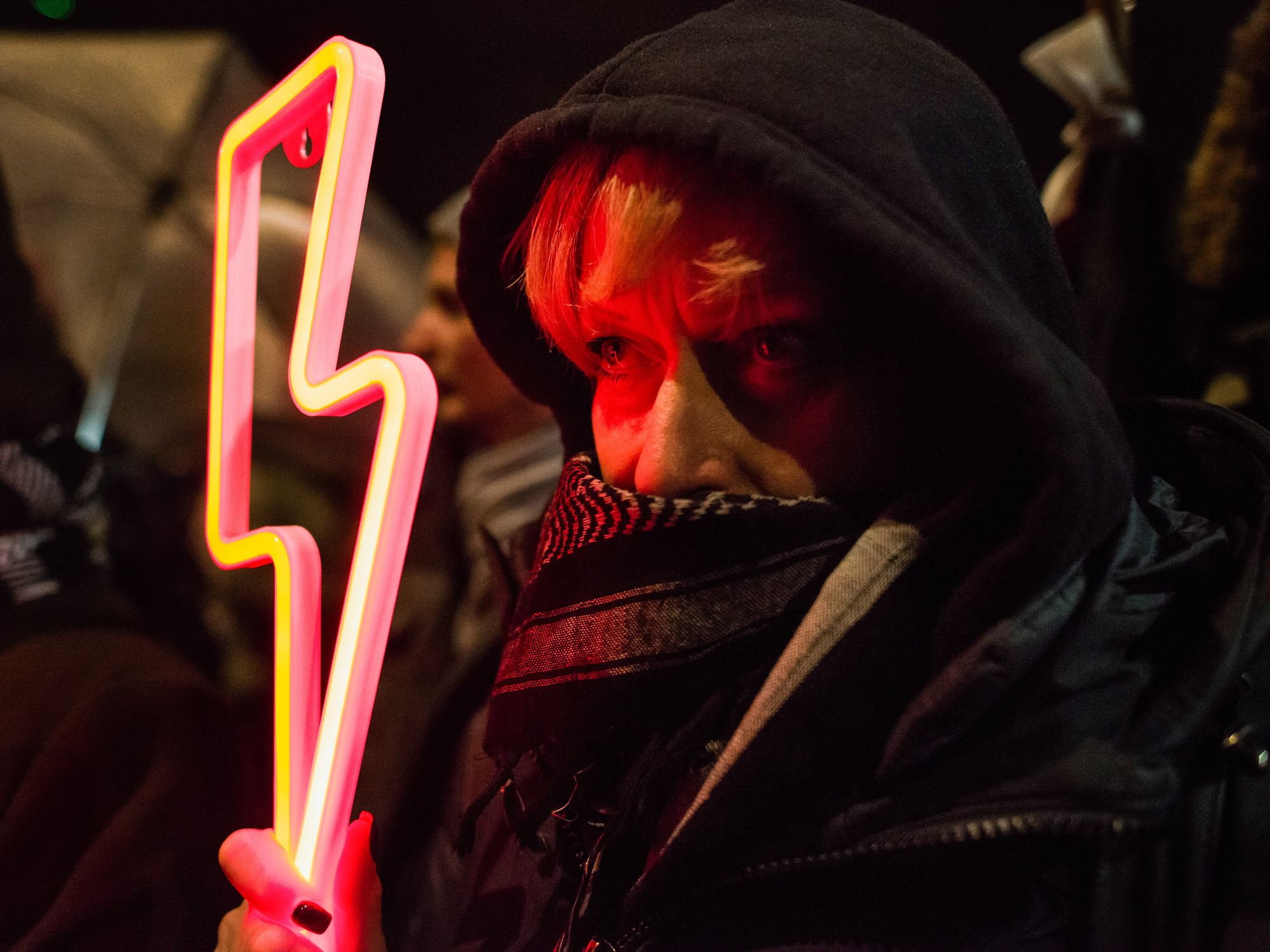 Photograph of a woman with her lower face covered holding a red lightning bolt - the symbol of the Women's Strike - during the demonstration outside Kaczynski's house.