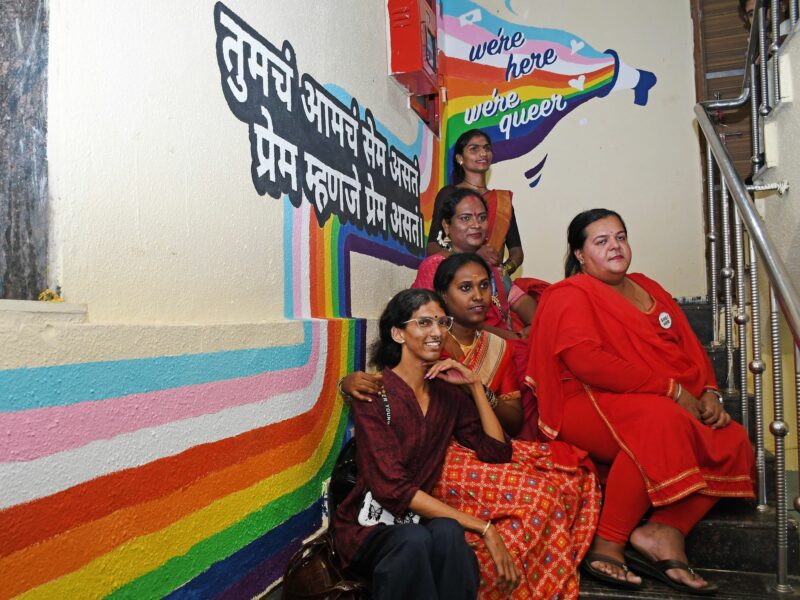 Photograph of five LGBTQ community members posing next to a rainbow mural outside Humsafar Trust.​