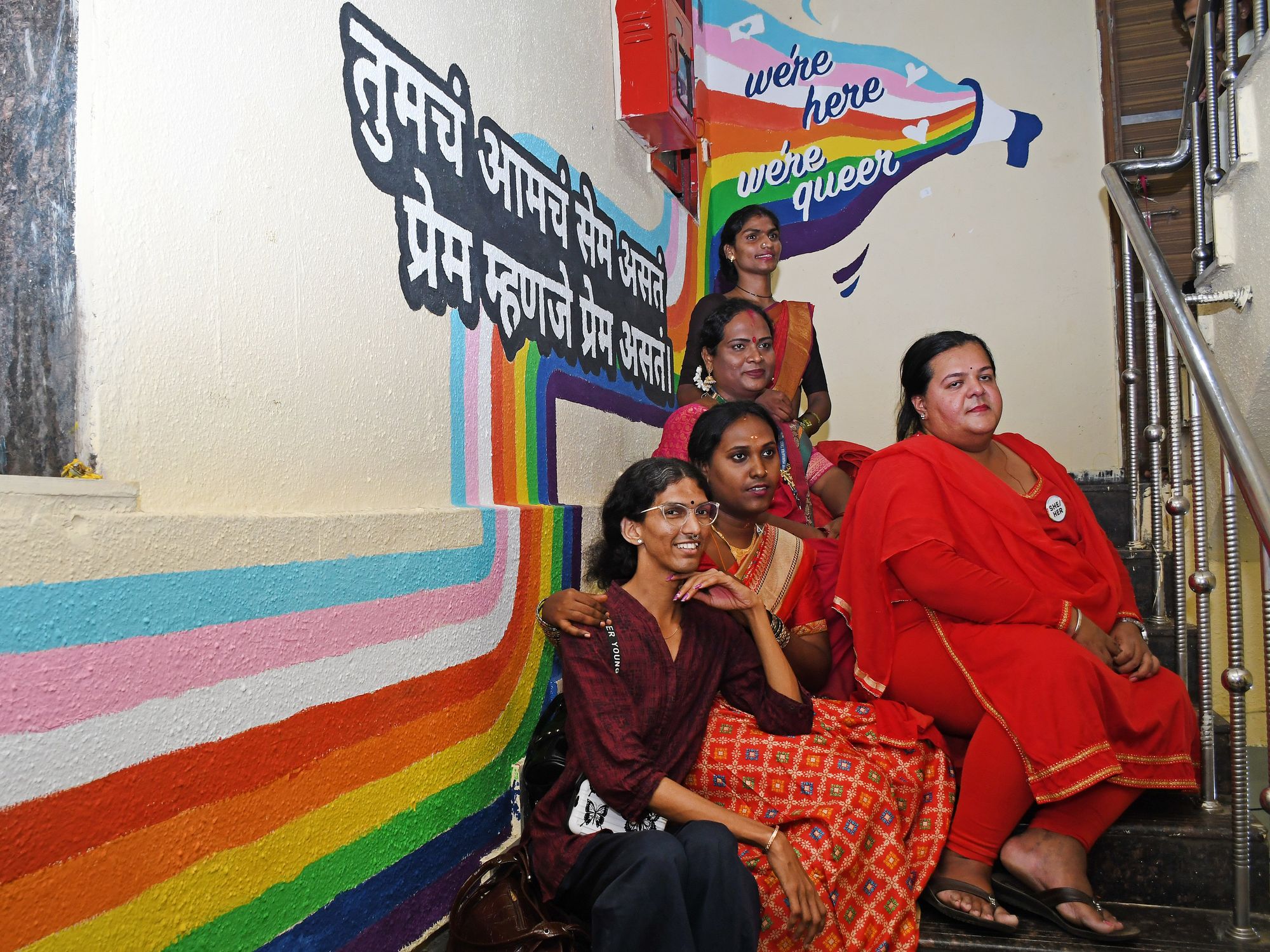 Photograph of five LGBTQ community members posing next to a rainbow mural outside Humsafar Trust.​