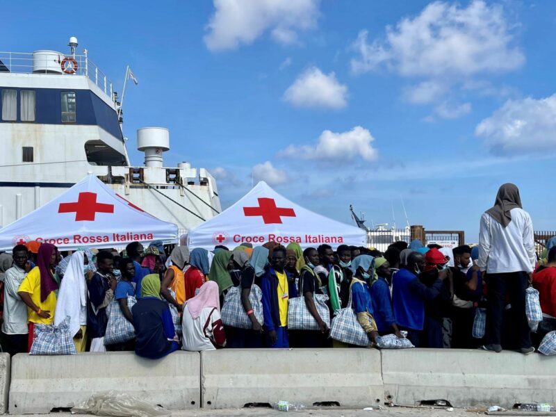 Photograph of migrants who have recently arrived to Lampedusa, standing in line as they wait to be transferred someplace else.