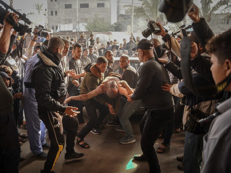 Photograph of Palestinians carry an injured man following the Israeli bombing on Khan Younis. They are surrounded by people and photographers.