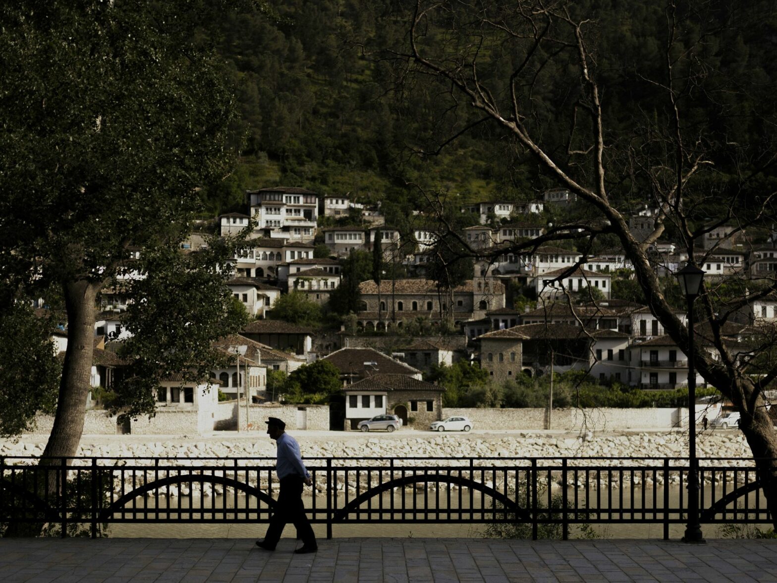 Photograph of the city view of the Albanian village of Berat. An old man walks along the river, surrounded by trees.