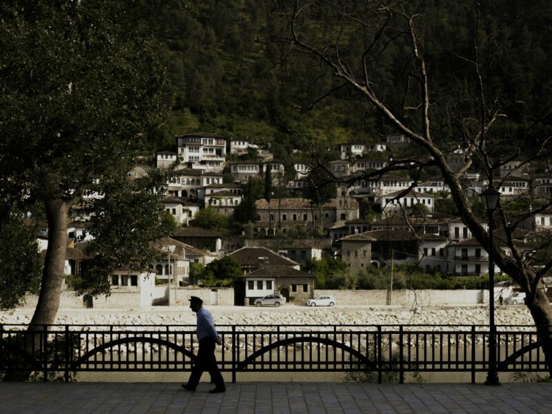 Photograph of the city view of the Albanian village of Berat. An old man walks along the river, surrounded by trees.