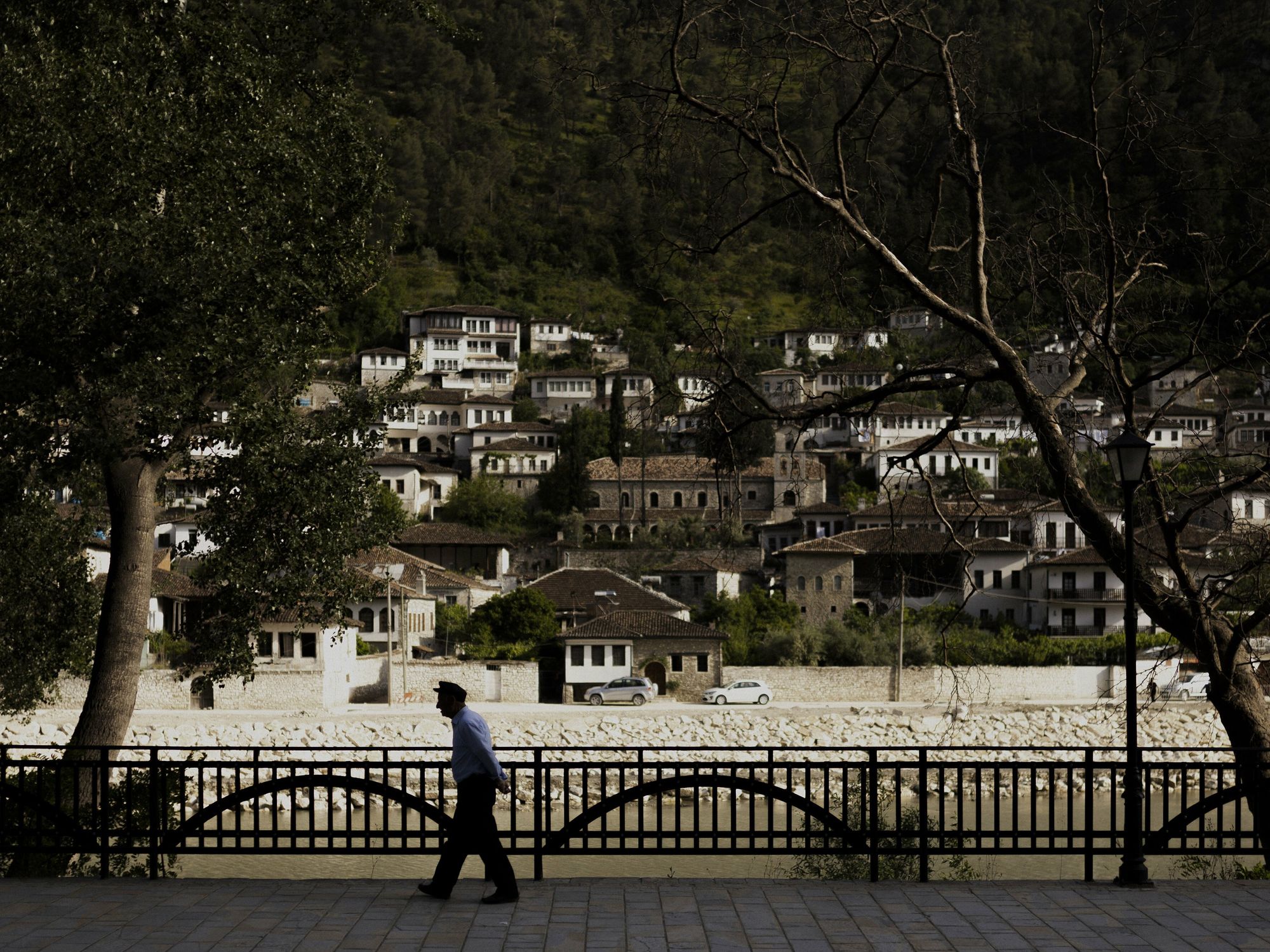 Photograph of the city view of the Albanian village of Berat. An old man walks along the river, surrounded by trees.