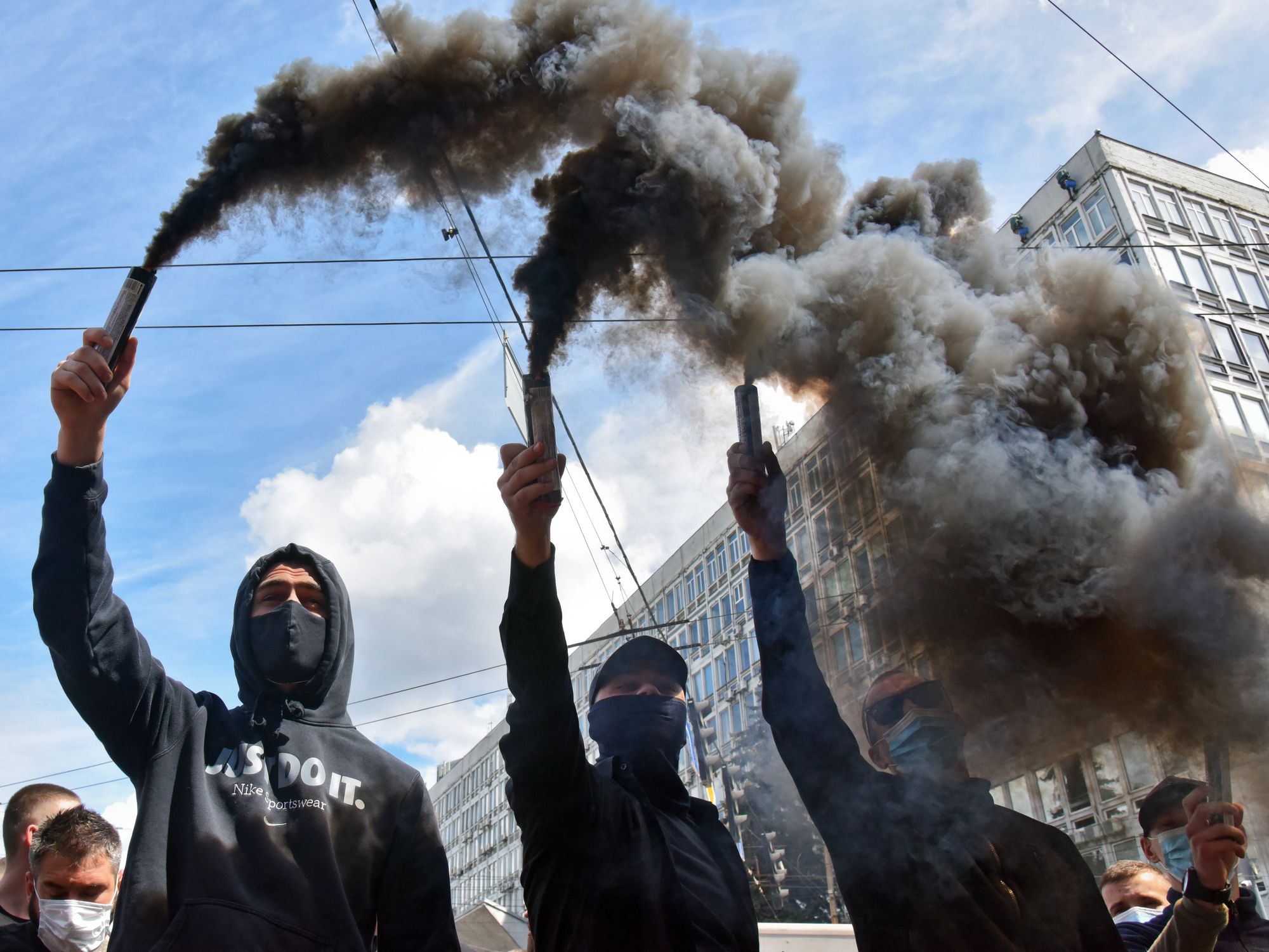 Photograph of three masked demonstrators holding black smoke lights.
