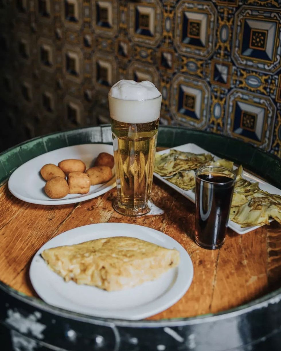 Plates of tapas including tortilla and croquetas are served alongside a beer
