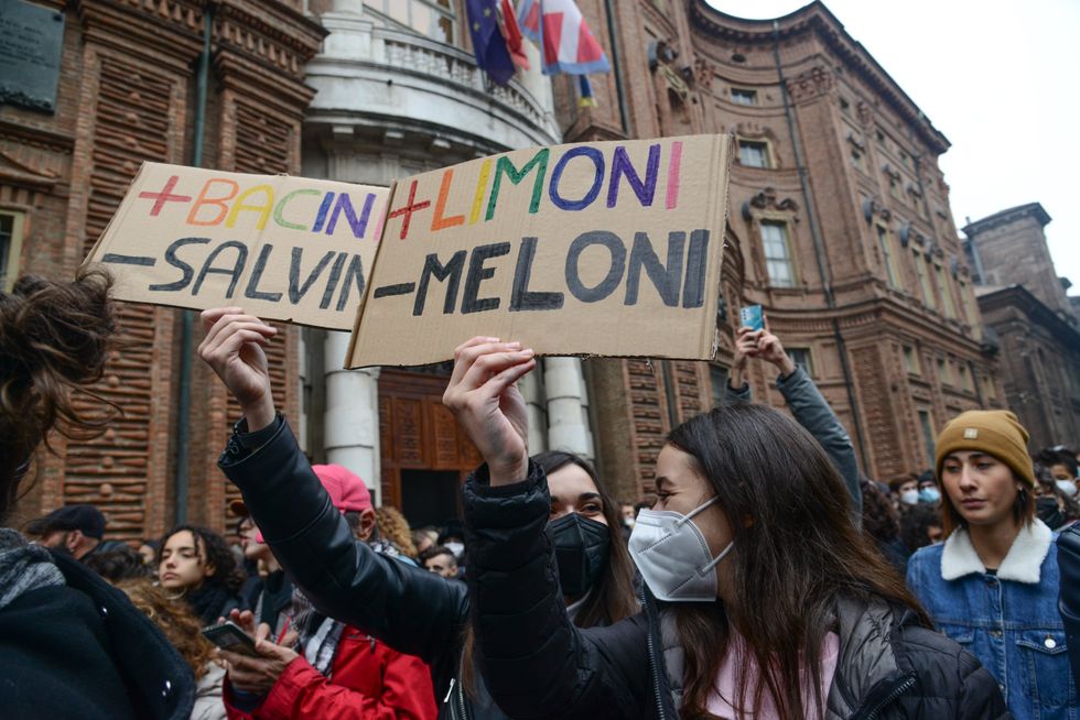 Protesters in the street, two women in medical masks hold signs saying "+ Bacini - Salvin" and "+ Limoni - Meloni"