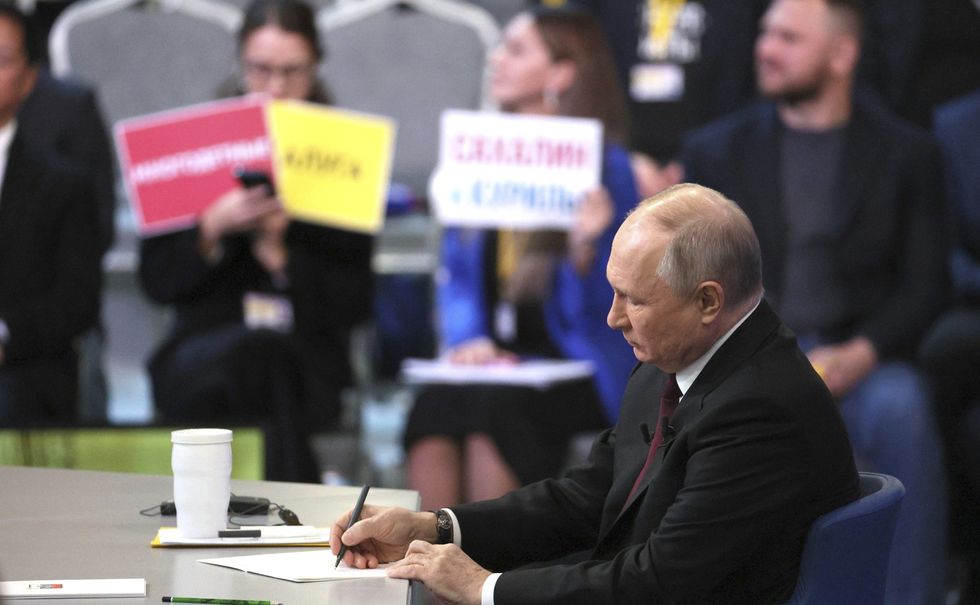 Russian President Vladmir Putin writing ona. piece of paper with journalist holding signs in the background.