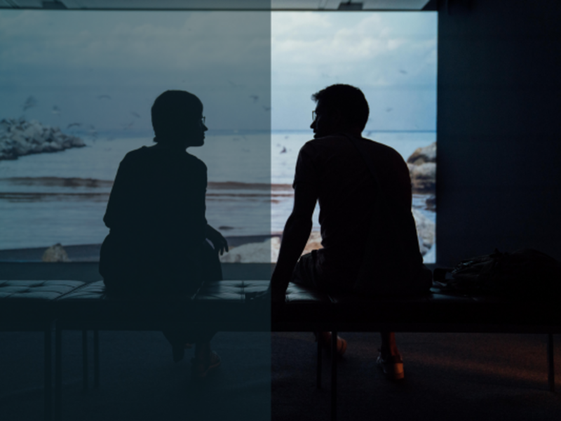 silhouette of man and woman sitting on ottoman as they look into each other's eyes. Behind them is a scene of the ocean.