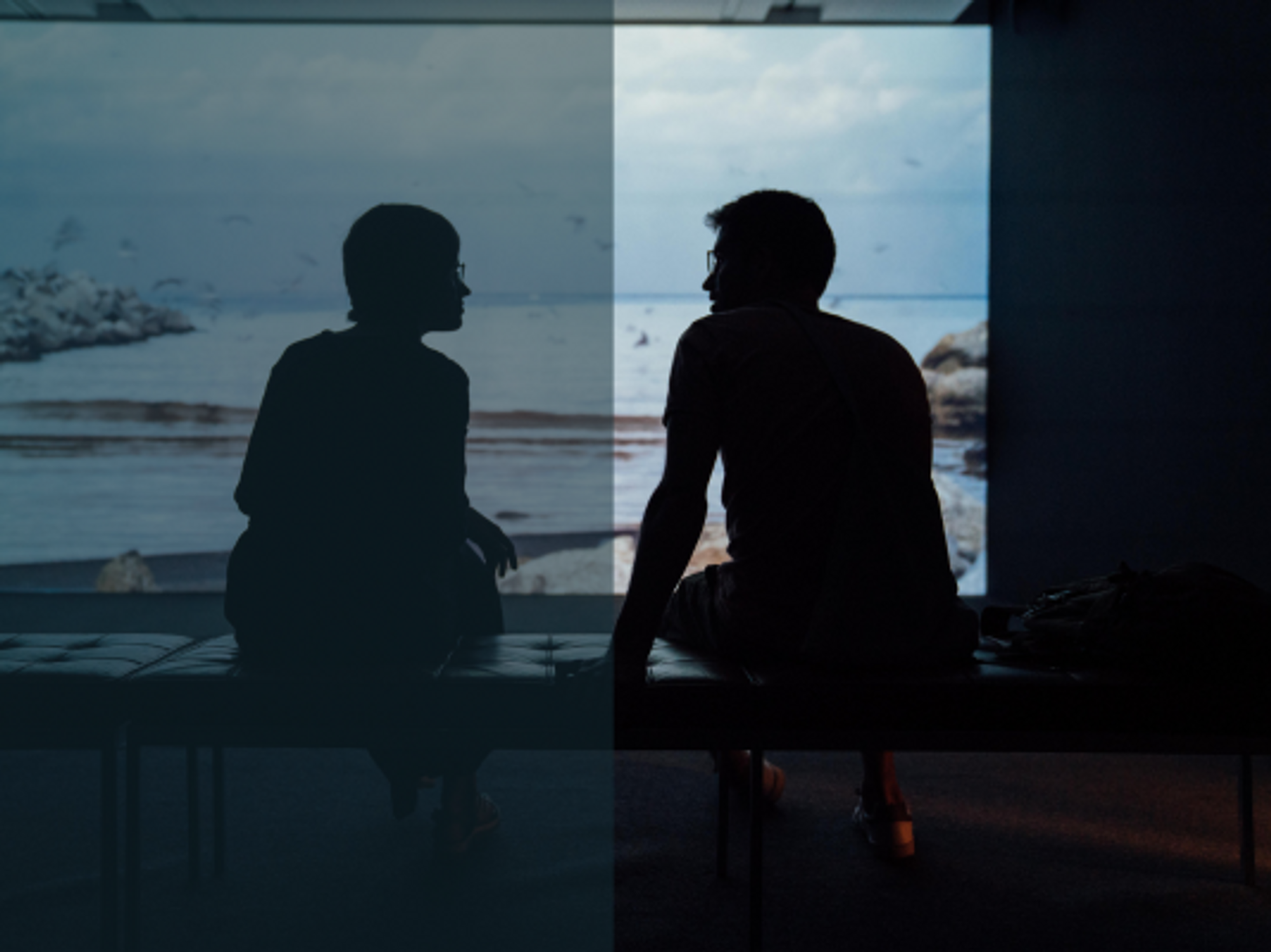 silhouette of man and woman sitting on ottoman as they look into each other's eyes. Behind them is a scene of the ocean.