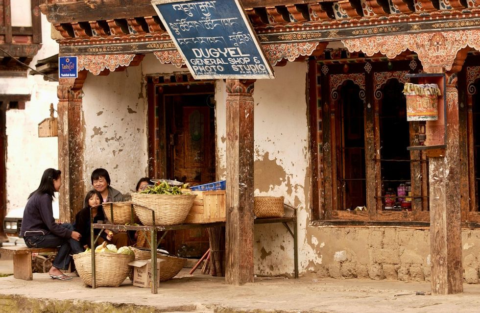 Street photo of a family of 4 sitting in front of a general shop & photoshop with baskets of fruits and vegetable on the table in front of them.