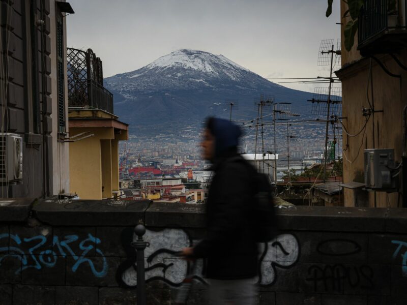 Street scene in Naples, with a person walking and in the background, snow-capped Vesuvius looming over Naples