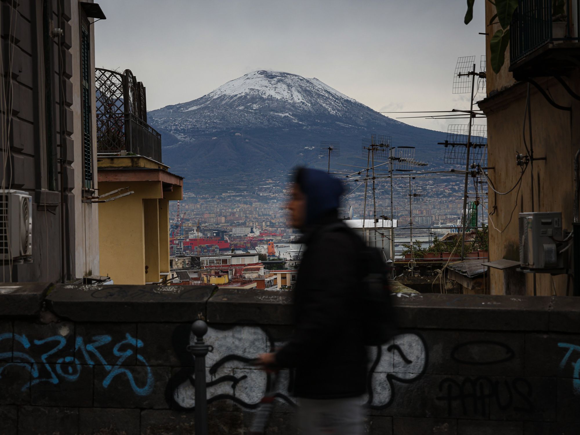 Street scene in Naples, with a person walking and in the background, snow-capped Vesuvius looming over Naples