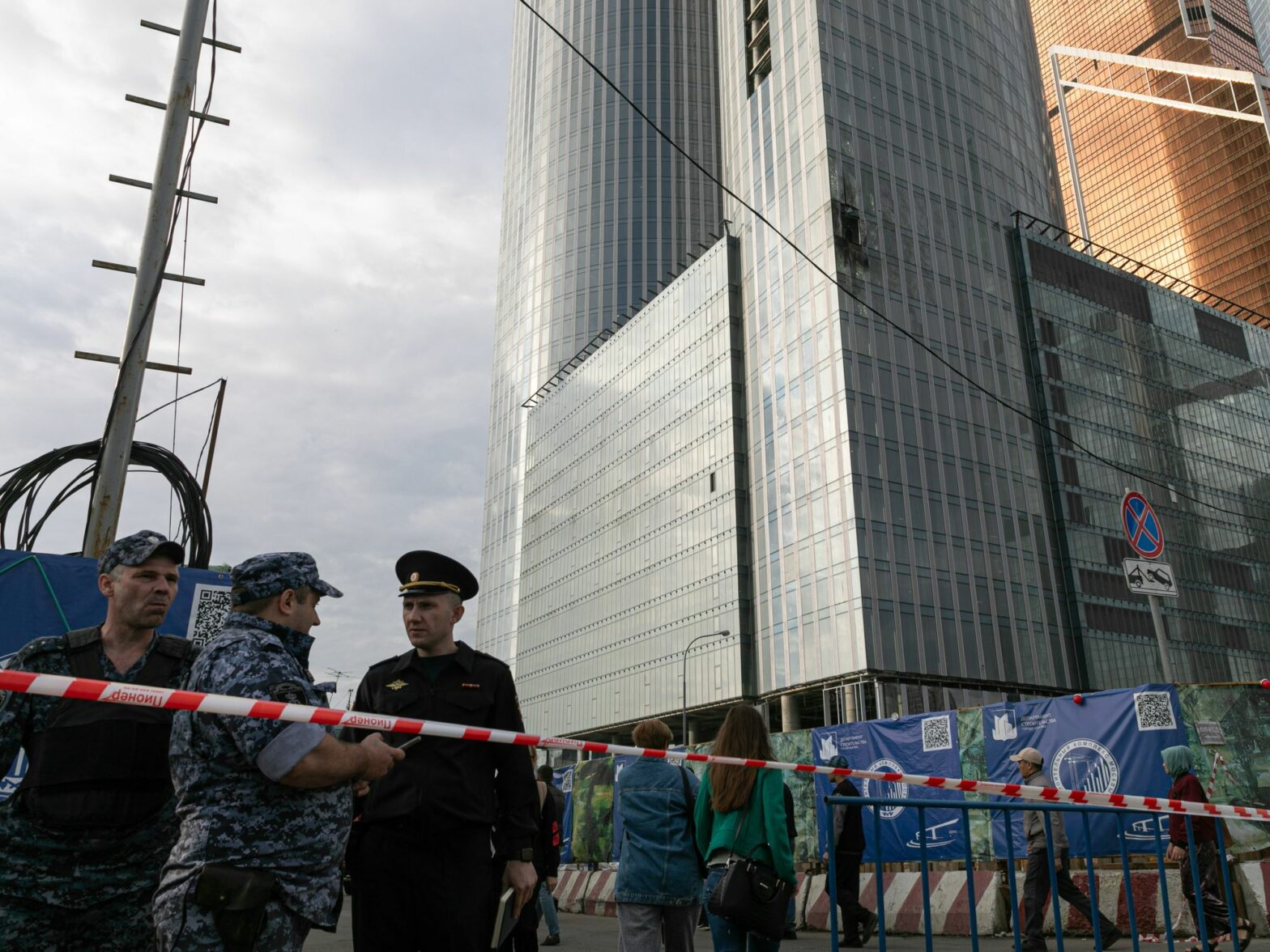 Tape guarded by russian offiials blocks off the street next to a drone damaged building