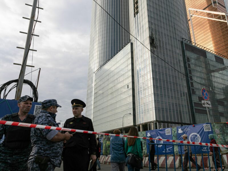 Tape guarded by russian offiials blocks off the street next to a drone damaged building