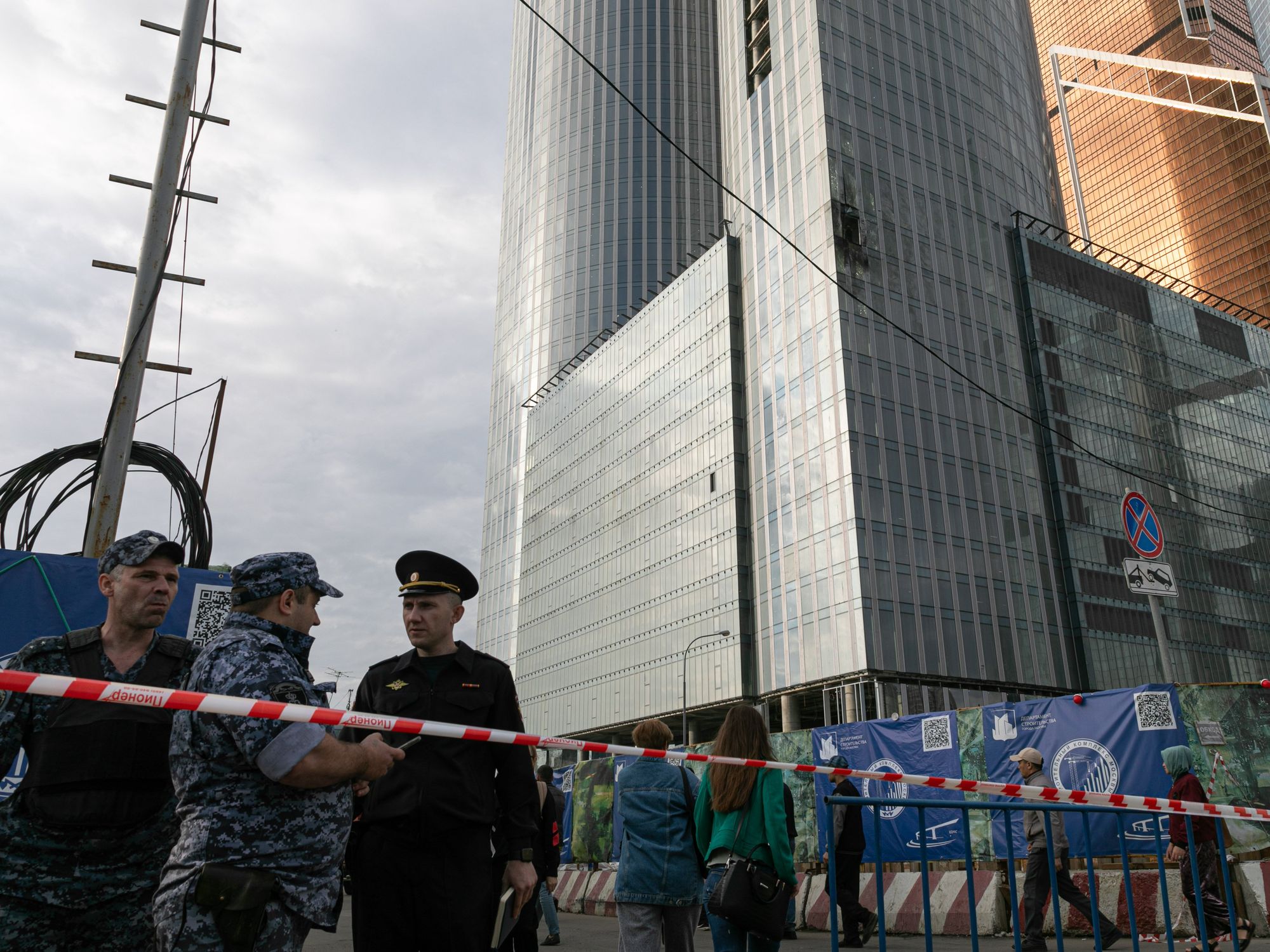 Tape guarded by russian offiials blocks off the street next to a drone damaged building