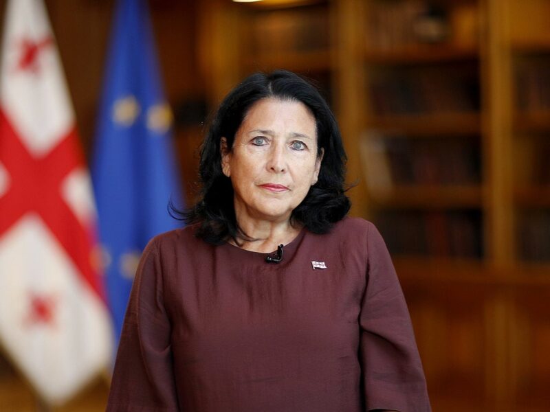This is a photograph of Georgian President Salome Zourabichvili, who stands and looks at the camera. In the background one can see a blurry Georgian flag next to the European Union flag.