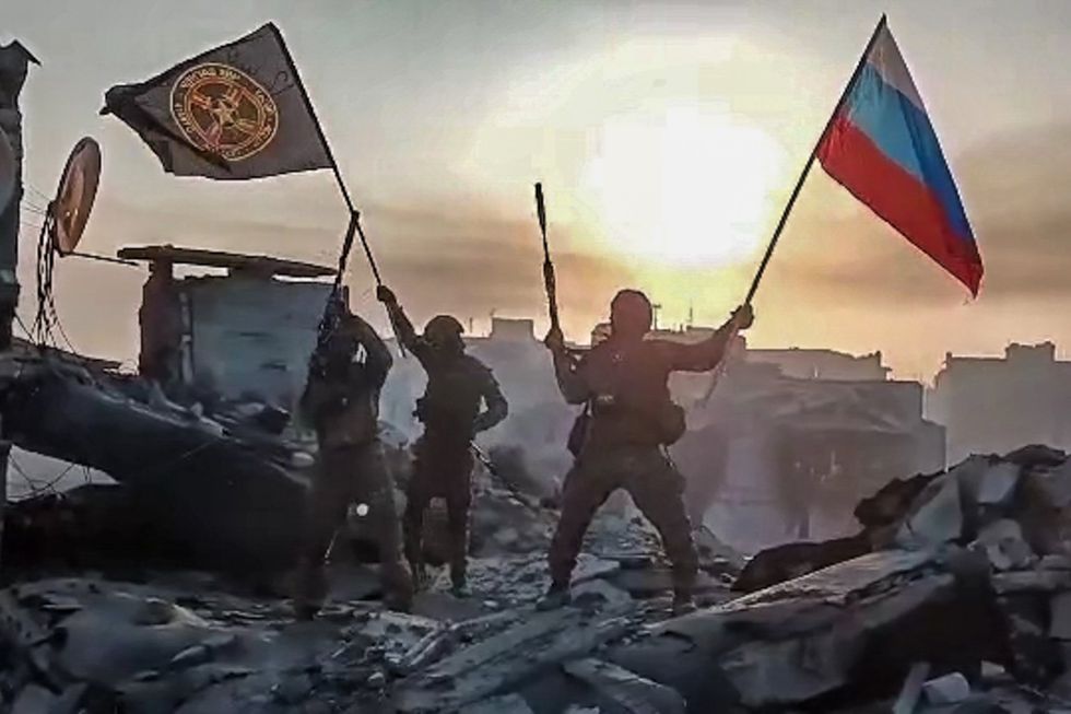 Three men stand on top of rubble, one is holding a Russian flag and one is holding the flag of Russia's Wagner Private Military Company.