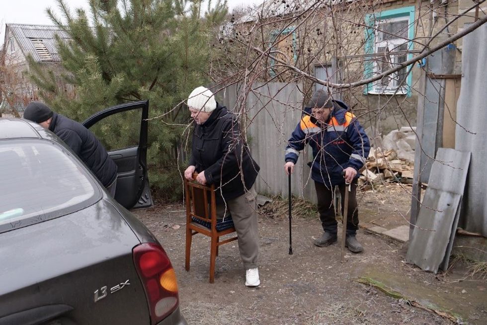 Three people load things into a car, with one person leaning on a chair and another person walking with a cane