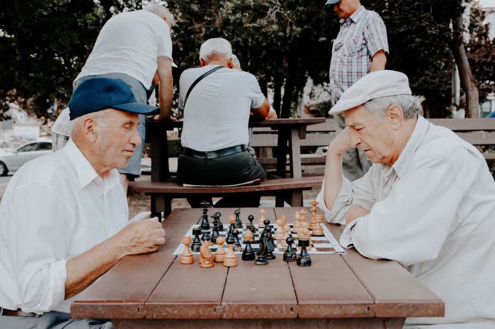 Two elderly men play chess at a park, behind them, more men play chess at a different table.