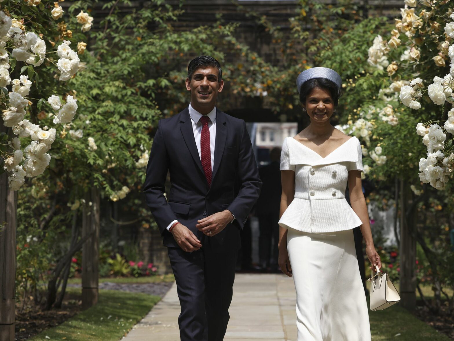 United Kingdom Prime Minister Rini Sunak and his wife walking down a sidewalk with flowers next to them. Their both dressed formally.