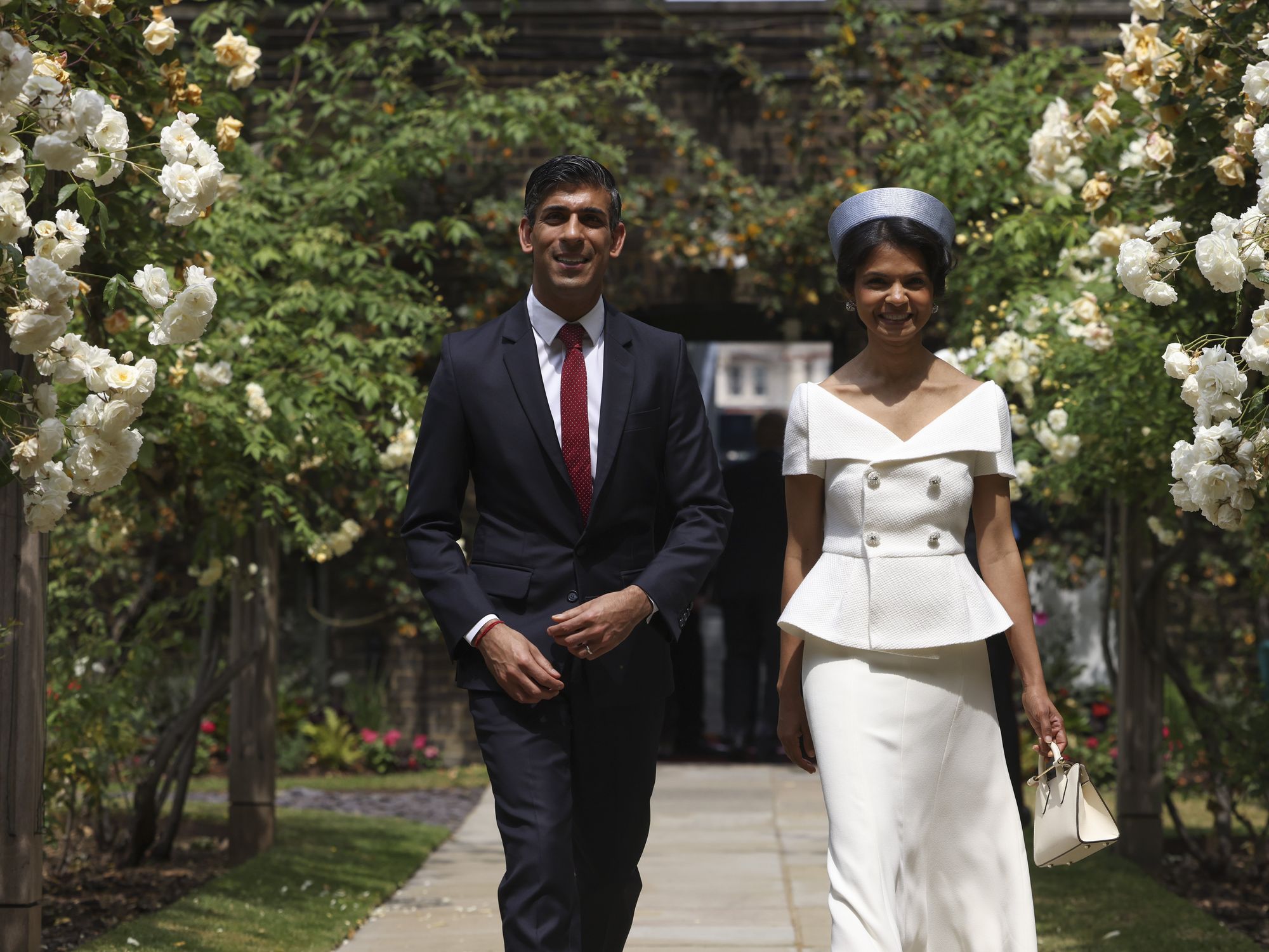 United Kingdom Prime Minister Rini Sunak and his wife walking down a sidewalk with flowers next to them. Their both dressed formally.