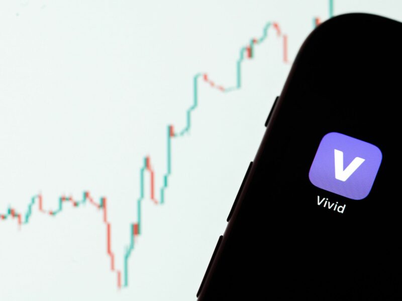 Vivid's app is seen on a display of an iPhone while the candlestick chart of a cryptocurrency is shown on a monitor in the background