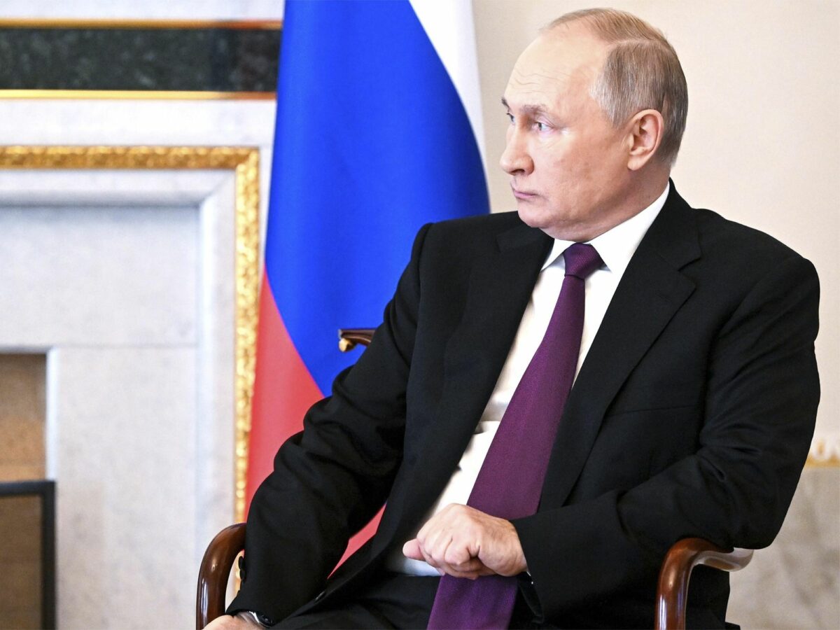 Vladimir Putin in a suit, sitting on a chair with russian flag in the background