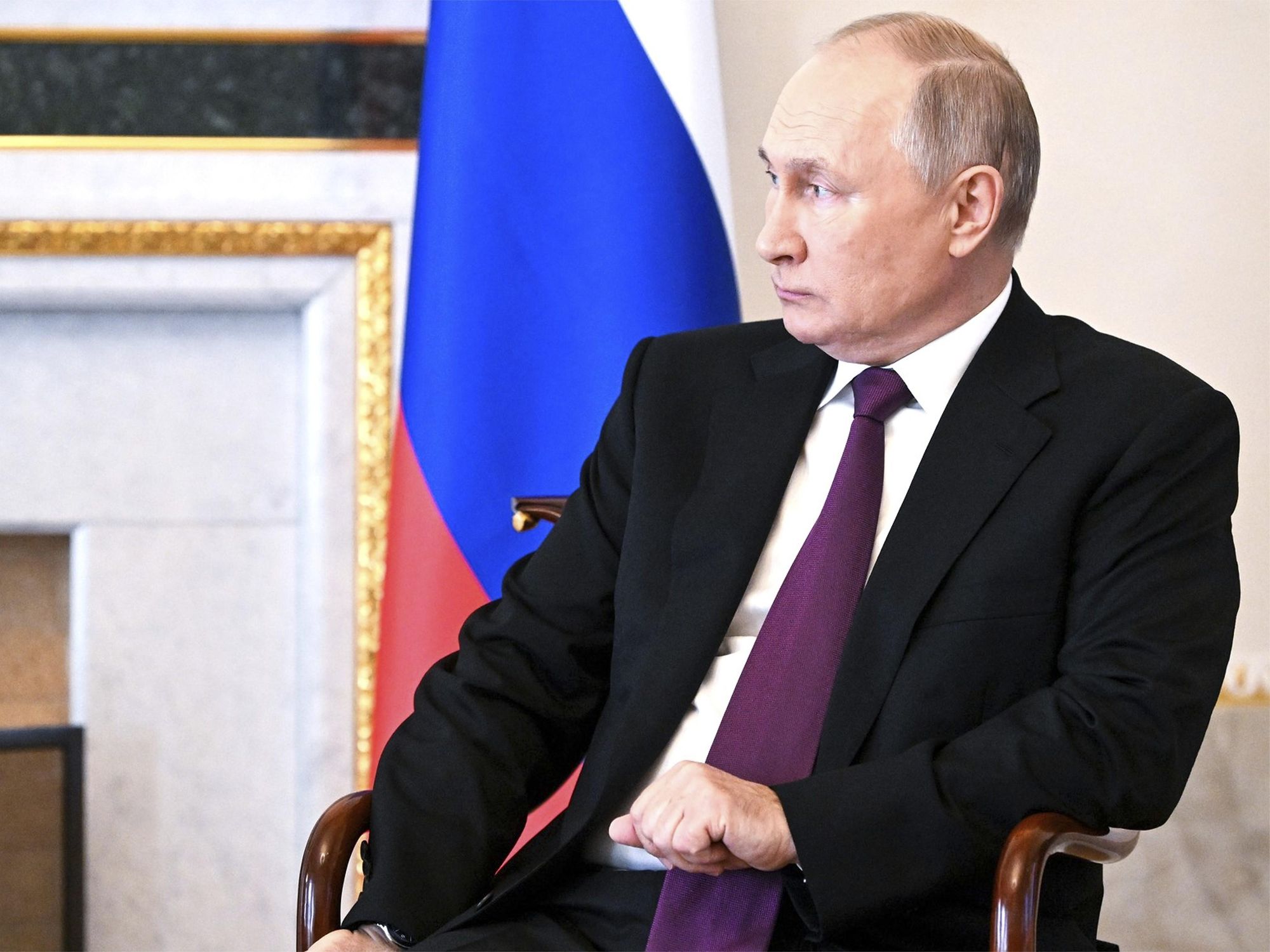 Vladimir Putin in a suit, sitting on a chair with russian flag in the background