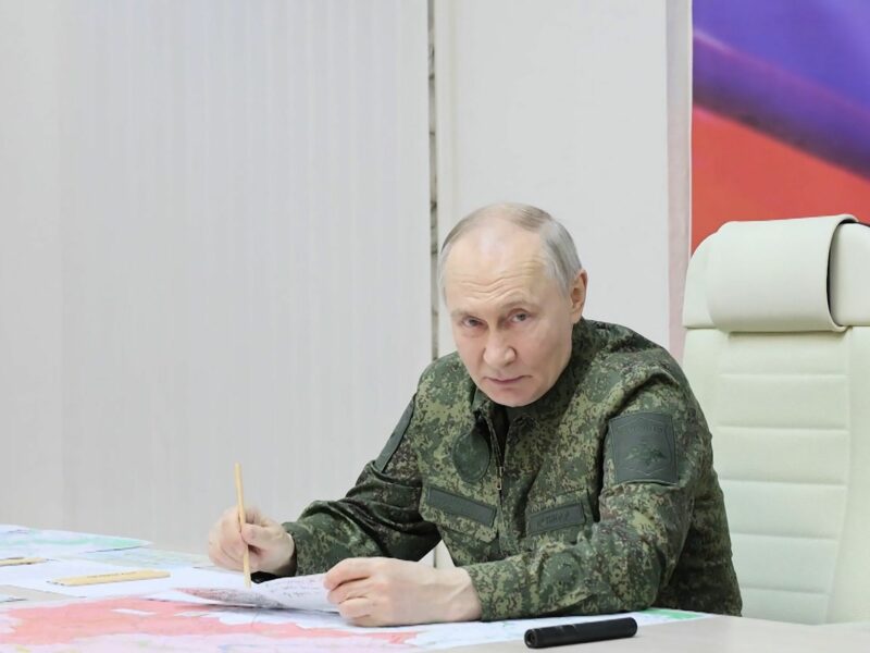 Vladimir Putin wears military fatigues and sits at a table with large colorful maps