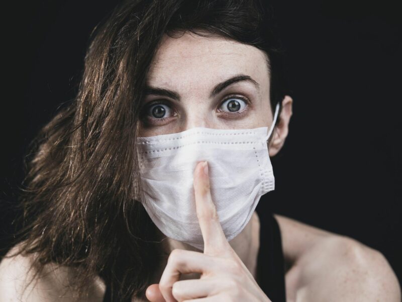woman wearing a surgical mask holding her finger in front of her mouth in a shush sign