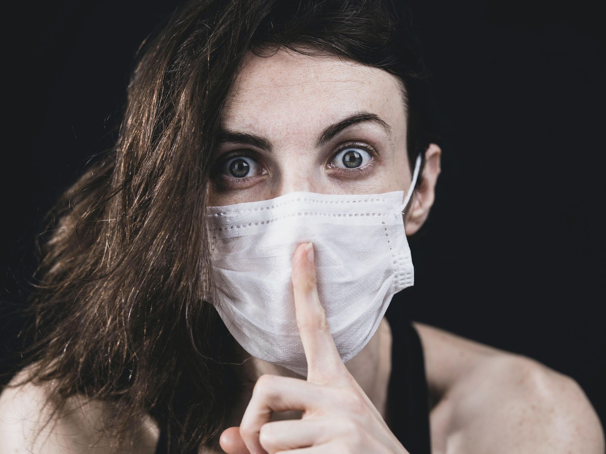 woman wearing a surgical mask holding her finger in front of her mouth in a shush sign