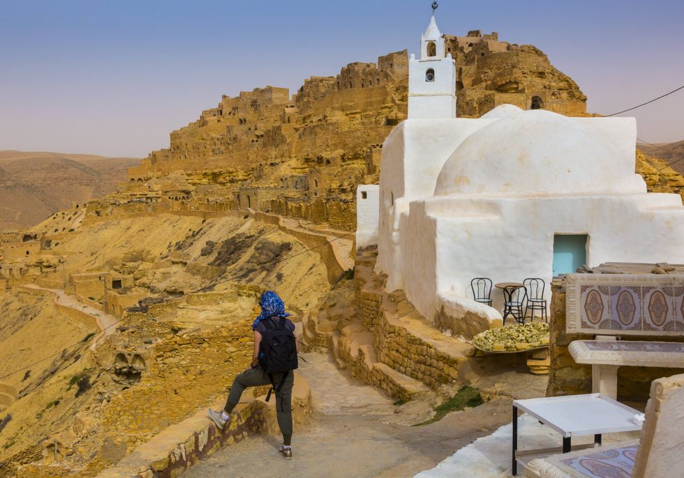women in head scarf standing at a balcony of a clay house, taking a look out at the ruins of a former ancient village