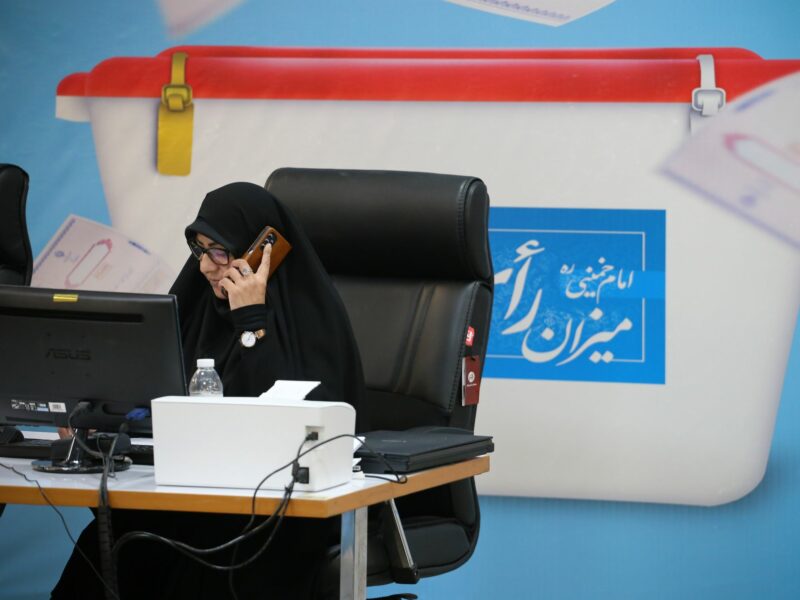 women in hijab in a chair with a phone to her ear, sitting behind her computer and in front of an image of an election box