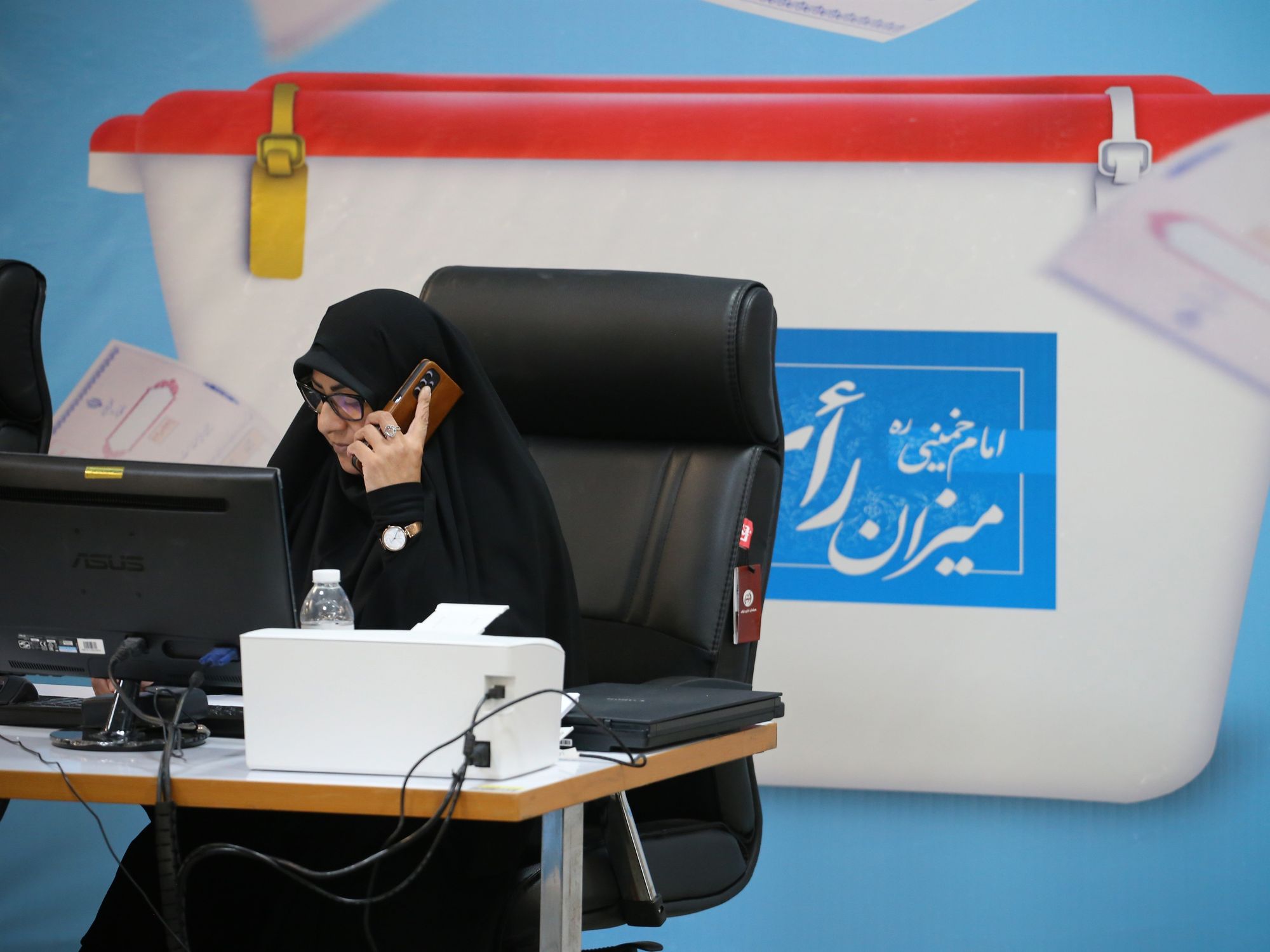 women in hijab in a chair with a phone to her ear, sitting behind her computer and in front of an image of an election box