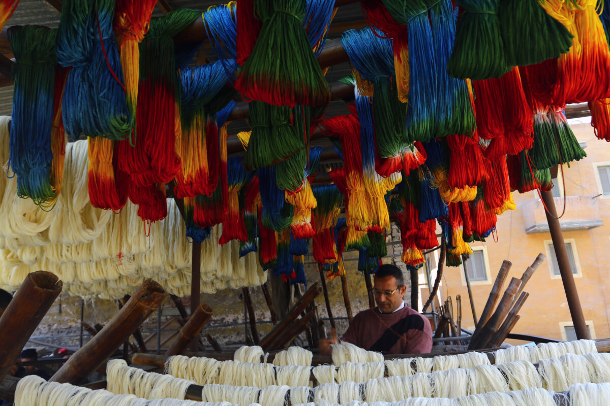 Hand-dyed yarn hangs to dry outside in rainbow patterns as a man walks through the hanging strings.