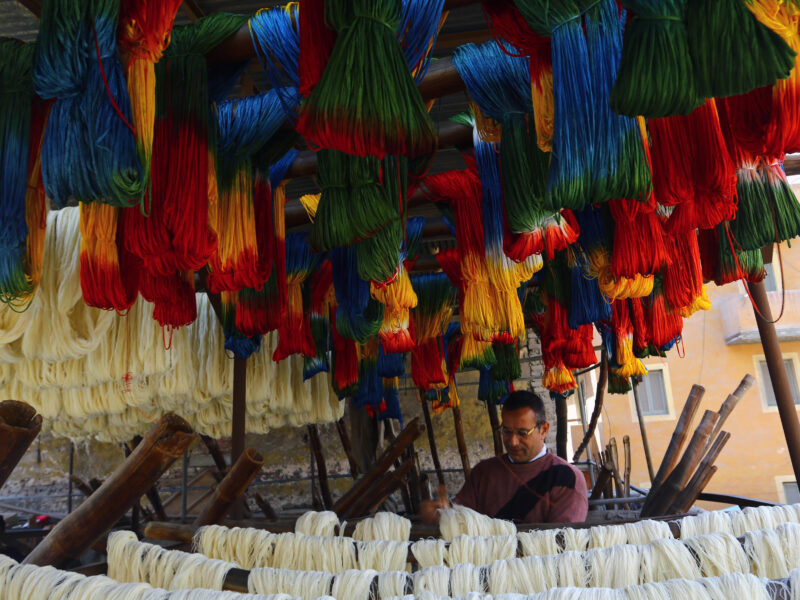 Hand-dyed yarn hangs to dry outside in rainbow patterns as a man walks through the hanging strings.