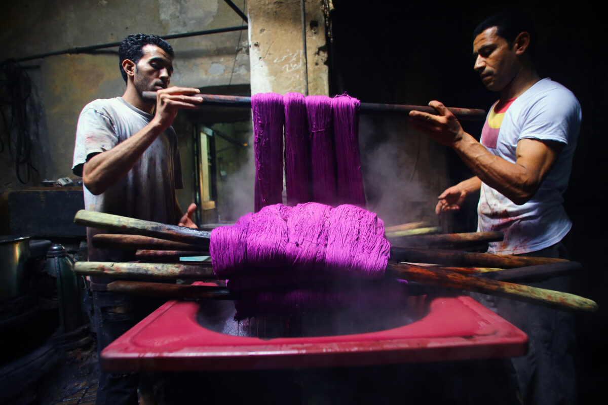 Two workers process purple yarn and pull it from a steaming ink bath.